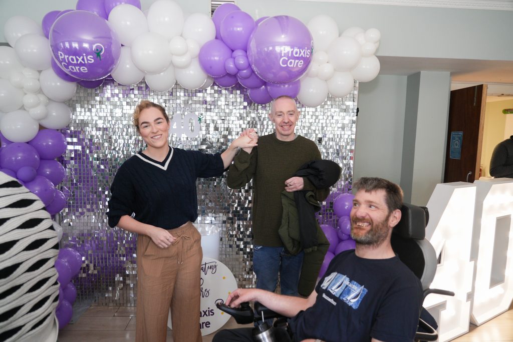 Two people stand holding hands with a man in a wheelchair beside them, all smiling in front of a sparkly feature wall with a border of purple and white balloons