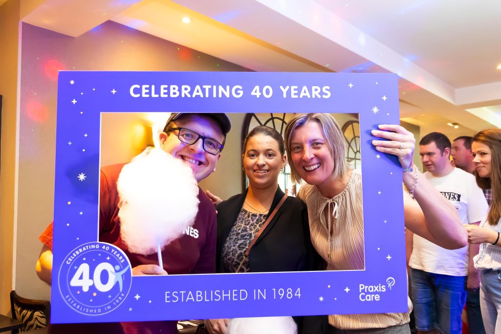 three people smile and pose in a prop frame that says Celebrating 40 Years. One person is holding candy floss on a stick.