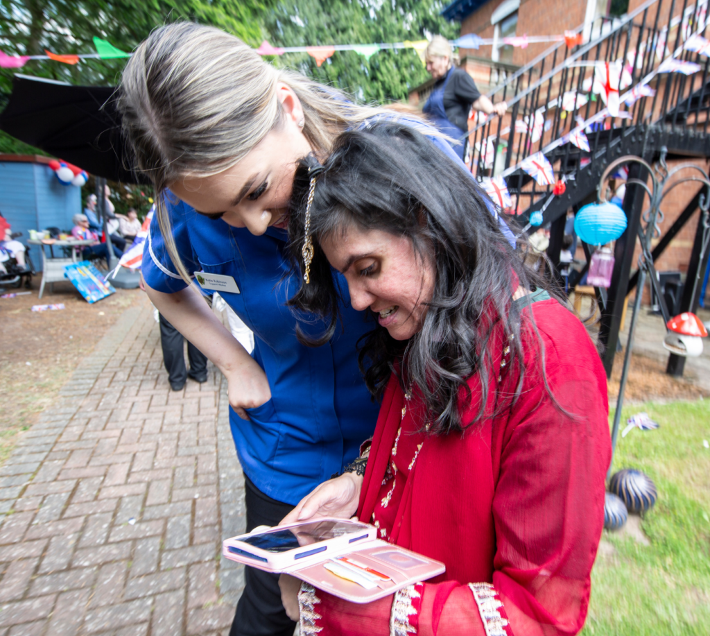 Blonde nurse in blue uniform assisting woman with dark hair in red top outdoors at nurse led service, looking at phone with smiles.