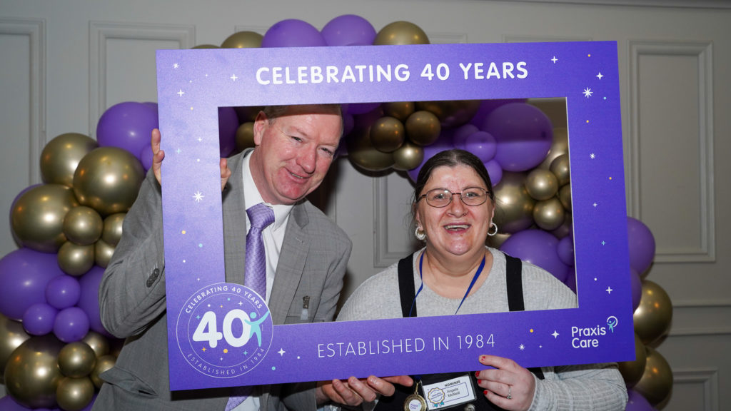 Man and woman posing holding a Praxis purple frame. Purple and gold balloons in the background.